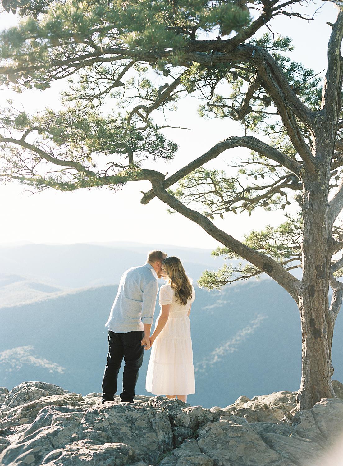 Groom leans in to whisper something in brides ear as they over look The Blue Ridge Mountains during their engagement session at Ravens Roost.